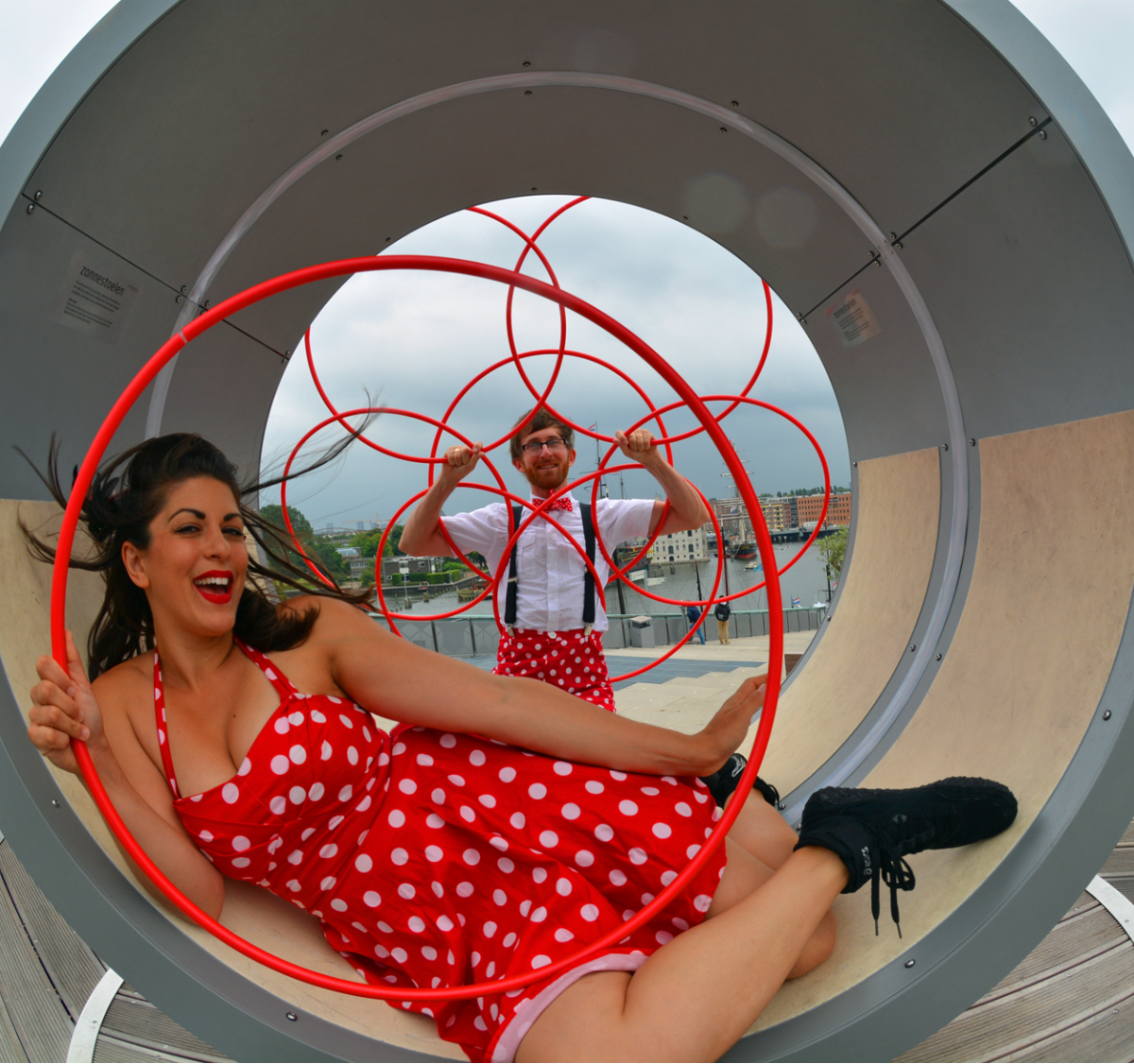 Woman in a red polka dot dress sitting inside a large circular sculpture with a man standing outside with hula hoops.