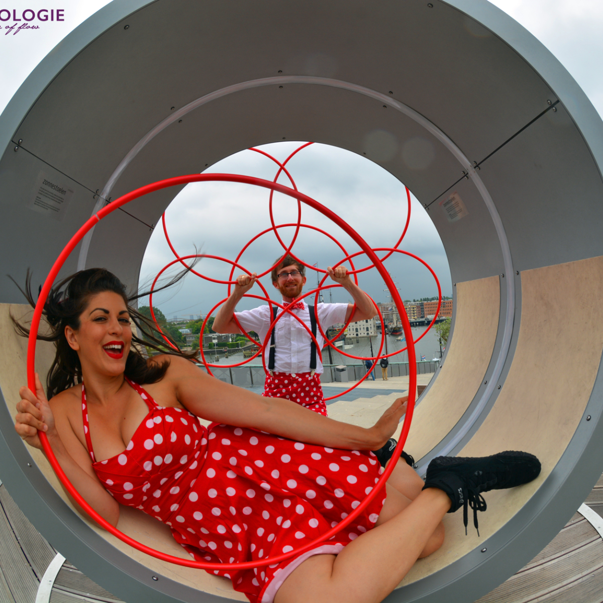 Woman in a red polka dot dress sitting inside a large circular sculpture with a man standing outside with hula hoops.